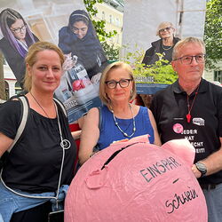 2 Frauen und ein Mann halten ein übergroßes Schwein auf dem Einsparschwein steht