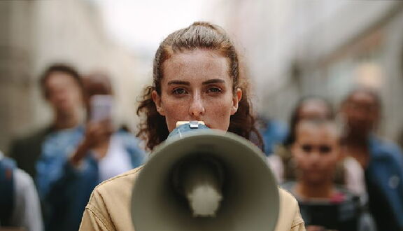 Eine Frau spricht in ein Megaphon und schaut dabei direkt in die Kamera.